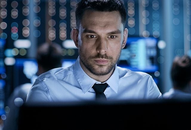 Close-up of a Professional Technical Controller Sitting at His Desk with Multiple Displays Before Him. In the Background His Colleagues Working in System Control Center.