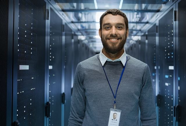 Portrait of a Curios, Positive and Smiling IT Engineer Standing in the Middle of a Large Data Center Server Room.