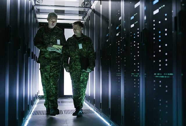 Two Military Men Walking in Data Center Corridor. One Uses Tablet Computer, They Have Discussion. Rows of Working Data Servers by their Sides.